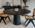 Flow round dining table in charcoal oak finish with black cylindrical base, featuring a sleek, modern design and subtle edge damage.